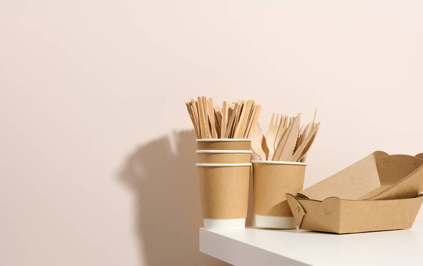 бумажные картонные коричневые тарелки и чашки, деревянные вилки и ножи на белом столе, бежевый фон. Экологичная посуда, нулевые отходы - Фото, изображение