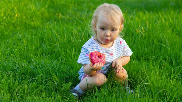 Baby girl is resting on the lawn on a hot summer day and eating a sweet donut. - Photo, image