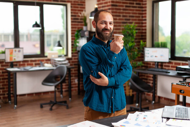 Portrét usměvavého podnikatele držícího šálek kávy stojící v kanceláři spouštěcí společnosti - Fotografie, Obrázek