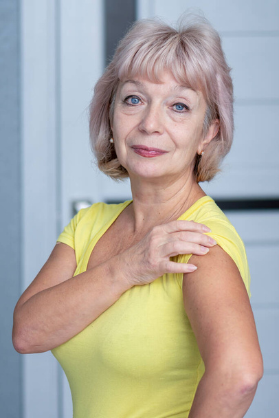 Μια όμορφη ηλικιωμένη γυναίκα 60-65 ετών δείχνει γυμνό ώμο της, ίσως αυτό είναι ένα σημάδι του εμβολιασμού, ένα μέρος για το κείμενο ή την εικόνα σας. - Φωτογραφία, εικόνα