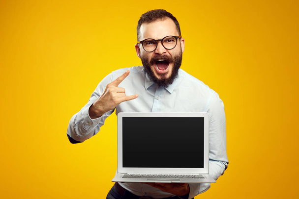Young caucasian male showing laptop screen and rock sign over yellow wall - Φωτογραφία, εικόνα
