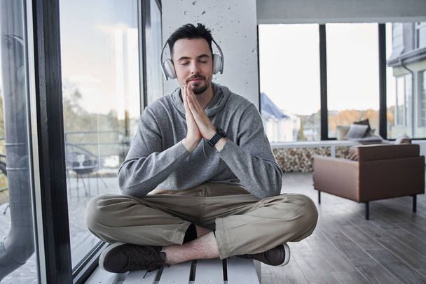 Man wearing headphones sitting with closed eyes at the lotus pose at the windowsill - Photo, image
