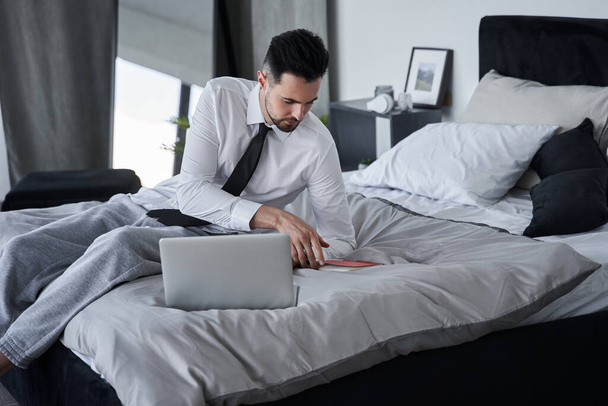 Man laying at the bed and reading something at his notebook while preparing to the work - Foto, Bild