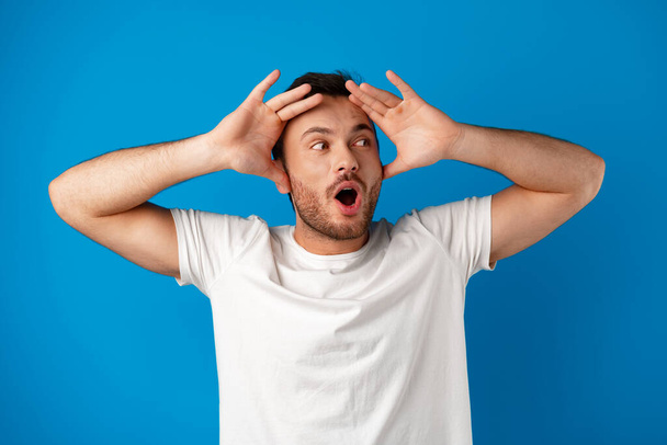 Portrait of a confused young man holding hands on his head over blue background - Photo, Image