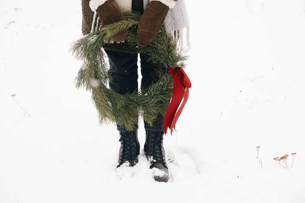 Vacanze invernali in campagna. Elegante donna con ghirlanda di Natale nel parco invernale. Giovane femmina in guanti accoglienti e cappotto tenendo ghirlanda di Natale con fiocco rosso nel campo invernale innevato, vista ritagliata - Foto, immagini