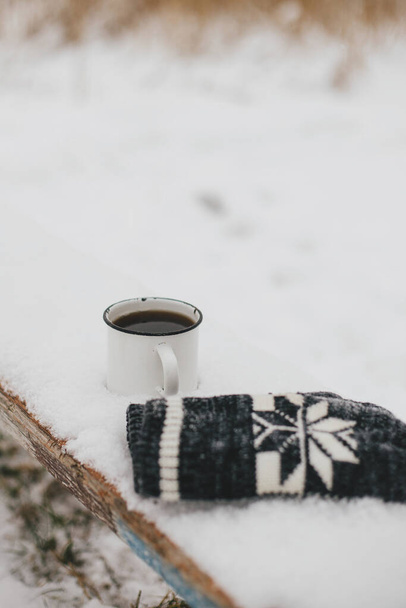 Warm cup of tea and cozy knitted gloves in snow on wooden bench in winter park. Hiking and traveling in cold season. Wanderlust. Space for text. Winter time - Photo, Image