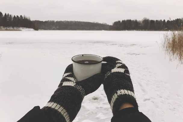 Las manos en guantes acogedores que sostienen la taza de té caliente en el fondo del lago de nieve en invierno. Senderismo y viajes en temporada fría de invierno. Un trago caliente. Espacio para el texto. Wanderlust - Foto, imagen
