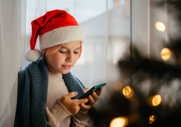 Junges glückliches Teenie-Mädchen mit roter Weihnachtsmütze mit Handy und Weihnachtsbaum im Vordergrund - Foto, Bild