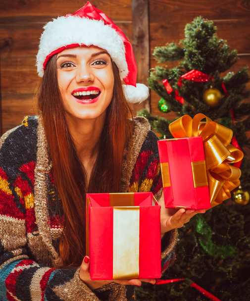 Caucasian Brunette Lady with a Wide Smile in a Casual Sweater and a Santa Hat Holds an Open Red Gift Box in her Hands Next to the Christmas Tree. Christmas Gifts, Christmas Mood. Wood Background - Zdjęcie, obraz