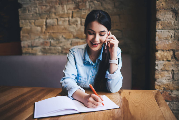 Happy female freelancer looking down while sitting at table writing notes in notebook and having conversation on mobile phone during workday - Foto, Imagem