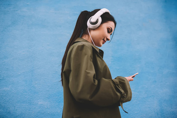 Side view of cheerful young lady with dark hair in coat smiling and browsing mobile phone while listening to music with headphones against blue background - Foto, Imagen