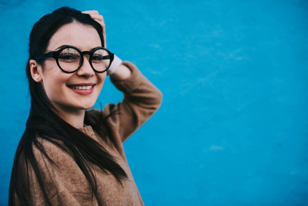 Happy female in casual sweater with long dark hair and eyeglasses looking at camera while leaning on blue wall and putting hand on head - Foto, afbeelding
