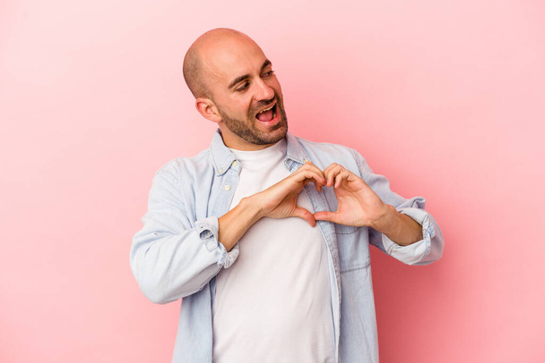 Jovem homem careca caucasiano isolado em fundo rosa sorrindo e mostrando uma forma de coração com as mãos. - Foto, Imagem