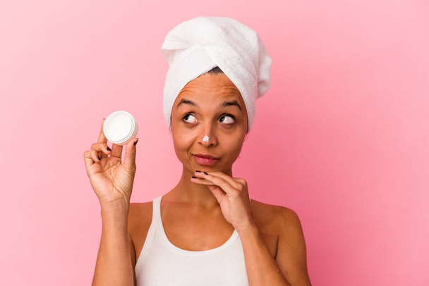 Young mixed race woman holding facial cream isolated on pink background looking sideways with doubtful and skeptical expression. - Photo, Image