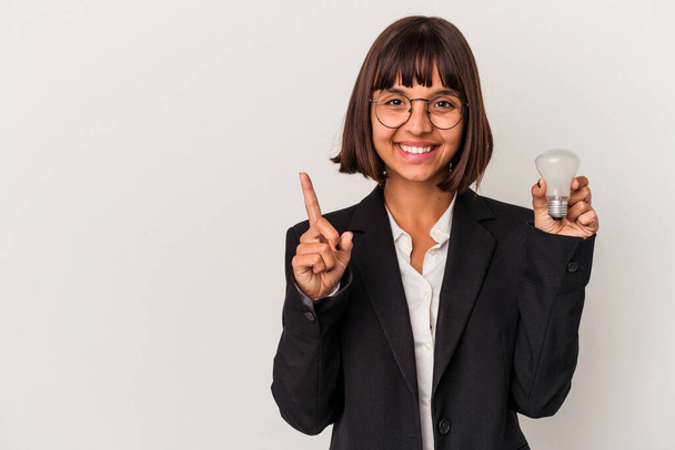 Jeune femme d'affaires métisse tenant une ampoule isolée sur fond blanc montrant numéro un avec doigt. - Photo, image