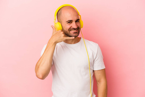 Молодой кавказский лысый мужчина слушает музыку на розовом фоне, показывая жест мобильного телефона с пальцами. - Фото, изображение