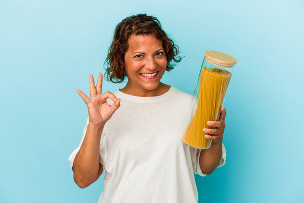 Middle age latin woman holding pasta jar isolated on blue background cheerful and confident showing ok gesture. - Photo, Image