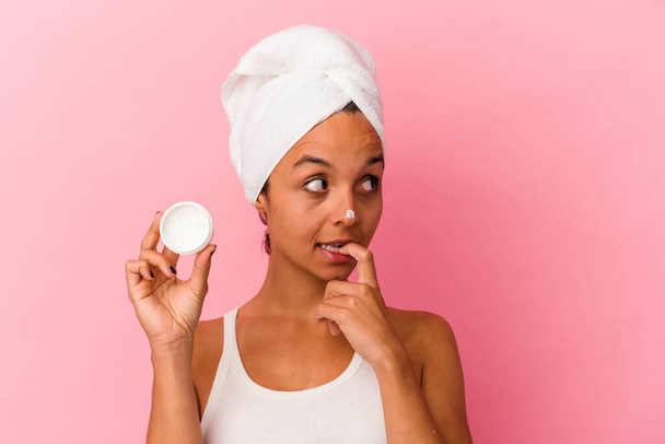 Young mixed race woman holding facial cream isolated on pink background relaxed thinking about something looking at a copy space. - Фото, изображение