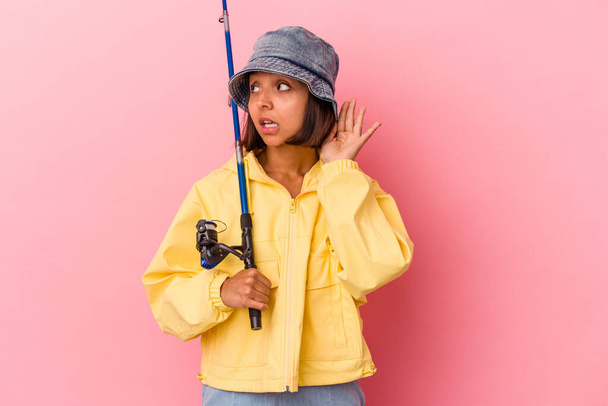 Joven mujer de raza mixta practicando la pesca aislada sobre fondo rosa tratando de escuchar un chisme. - Foto, imagen