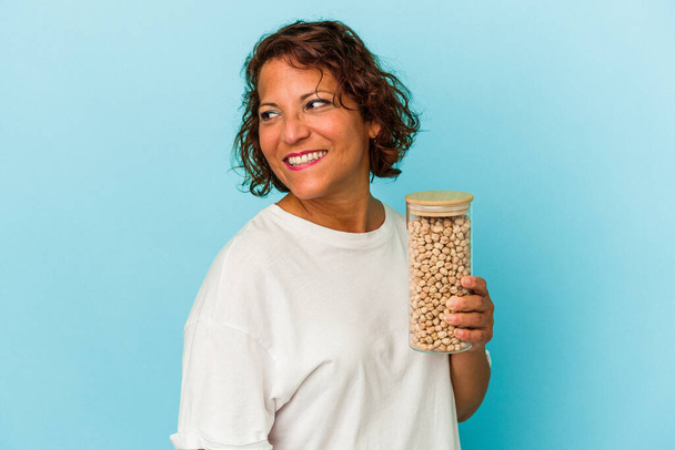 Middle age latin woman holding chickpeas jar isolated on blue background looks aside smiling, cheerful and pleasant. - 写真・画像