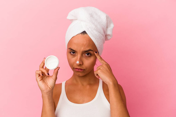 Young mixed race woman holding facial cream isolated on pink background pointing temple with finger, thinking, focused on a task. - Foto, Imagem