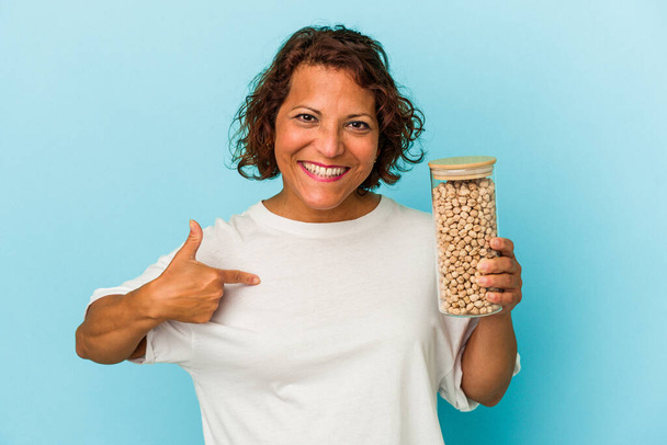 Middle age latin woman holding chickpeas jar isolated on blue background person pointing by hand to a shirt copy space, proud and confident - Photo, Image
