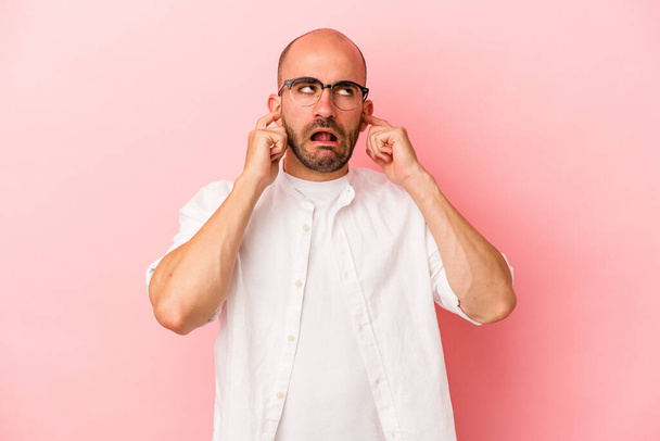 Jovem homem careca caucasiano isolado em fundo rosa cobrindo orelhas com os dedos, estressado e desesperado por um ambiente barulhento. - Foto, Imagem