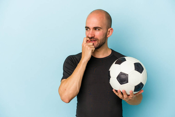 młody biznes łysy człowiek gra w piłkę nożną odizolowany na niebieskim tle zrelaksowany myślenie o czymś patrząc na przestrzeń kopii. - Zdjęcie, obraz
