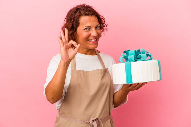 Mujer latina pastelera de mediana edad sosteniendo un pastel aislado sobre un fondo rosa alegre y confiado mostrando un gesto aceptable. - Foto, Imagen