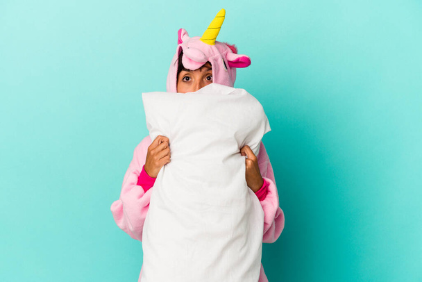 Young mixed race woman wearing a unicorn pajama holding a pillow isolated on blue background - Photo, Image