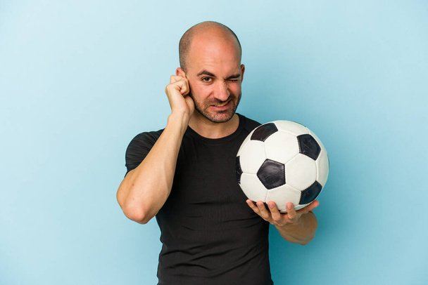 Young business bald man playing football isolated on blue background  covering ears with hands. - Foto, afbeelding