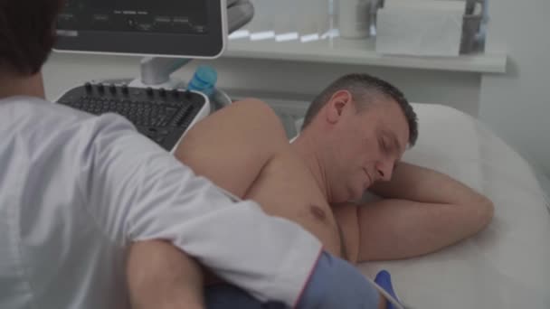 Doctor usando una máquina de ultrasonido para escanear el corazón de un paciente masculino. El cardiólogo hace de un paciente una prueba ecocardiográfica en una clínica. Paciente sometido a ecografía en el hospital - Imágenes, Vídeo