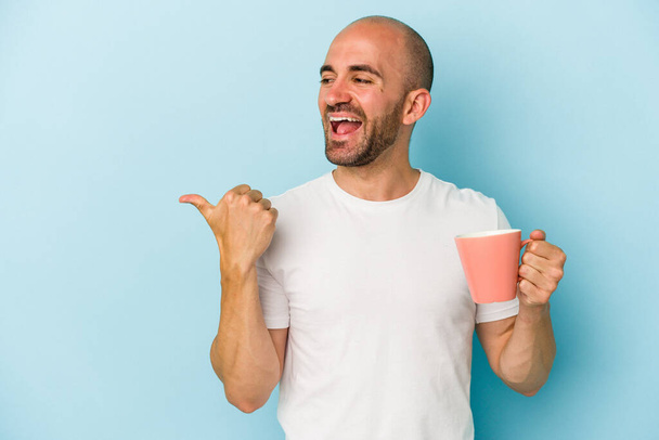 Young bald man holding a mug isolated on blue background  points with thumb finger away, laughing and carefree. - Foto, imagen