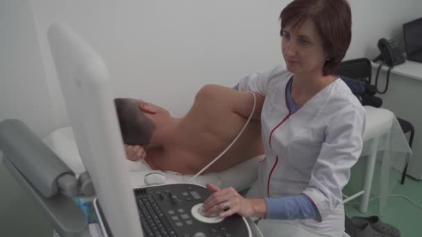 Lääkäri käyttää ultraäänikonetta miespotilaan sydämen skannaamiseen. Kardiologi tekee potilaasta kaikukardiografisen testin klinikalla. Potilas ultraäänitutkimuksessa sairaalassa - Materiaali, video