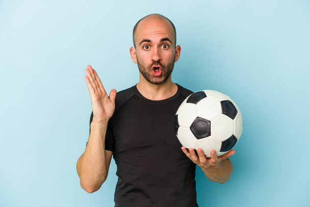 Young business bald man playing football isolated on blue background  surprised and shocked. - Valokuva, kuva