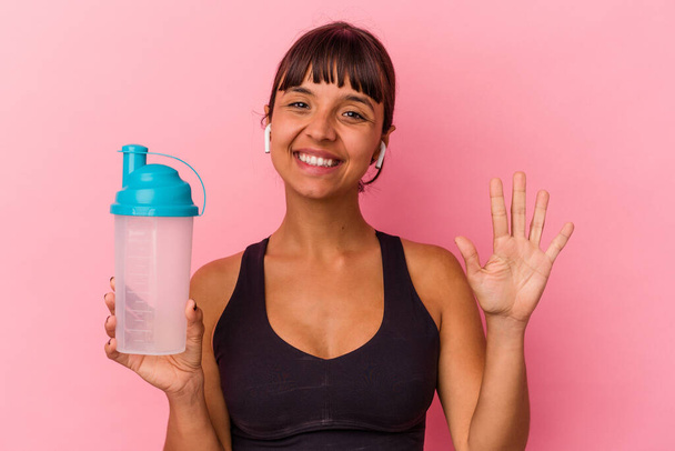 Giovane donna razza mista che beve frullato proteico isolato su sfondo rosa sorridente allegro mostrando il numero cinque con le dita. - Foto, immagini