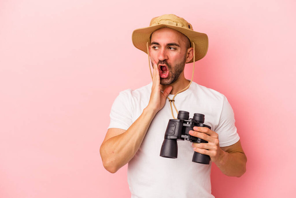 Young caucasian bald man holding binoculars isolated on pink background  is saying a secret hot braking news and looking aside - Photo, Image