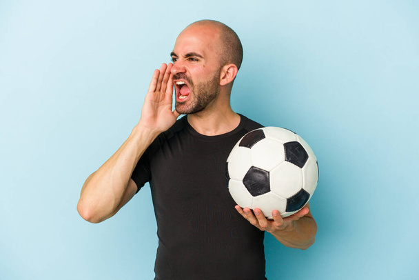 Νεαρός καραφλός παίκτης παίζει ποδόσφαιρο απομονωμένος σε μπλε φόντο φωνάζοντας και κρατώντας παλάμη κοντά άνοιξε το στόμα. - Φωτογραφία, εικόνα