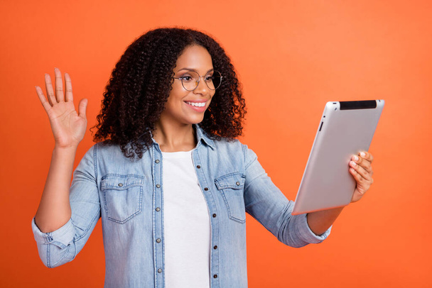 Photo of funny millennial brunette lady hold tablet wave wear jeans shirt isolated on orange color background - Foto, Imagem