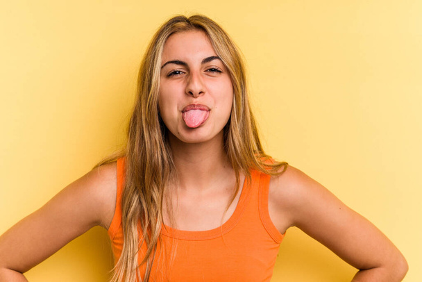 Young caucasian blonde woman isolated on yellow background  funny and friendly sticking out tongue. - Photo, Image