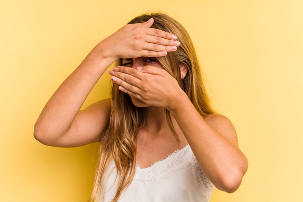 Nuori valkoihoinen blondi nainen eristetty keltaisella taustalla räpäyttää kameraa sormien kautta, hämmentynyt peittää kasvot. - Valokuva, kuva