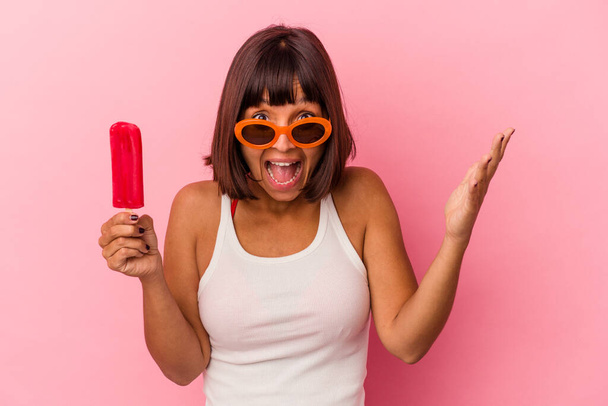 Young mixed race woman holding an ice cream isolated on blue background receiving a pleasant surprise, excited and raising hands. - Valokuva, kuva