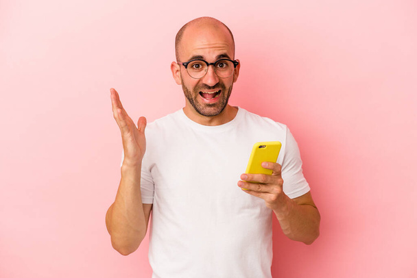 Fiatal kaukázusi kopasz férfi kezében mobiltelefon elszigetelt rózsaszín háttérrel kap egy kellemes meglepetés, izgatott és felemelt kezét. - Fotó, kép