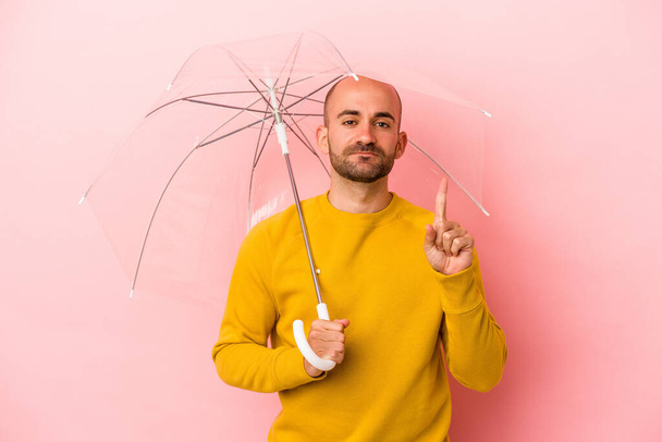 Young caucasian bald man holding umbrella isolated on pink background  showing number one with finger. - Photo, Image