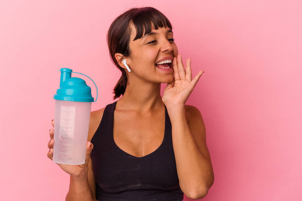 Joven mujer de raza mixta bebiendo batido de proteínas aislado sobre fondo rosa gritando y sosteniendo la palma cerca de la boca abierta. - Foto, Imagen