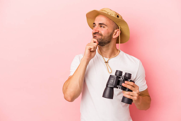 Young caucasian bald man holding binoculars isolated on pink background  looking sideways with doubtful and skeptical expression. - Photo, Image