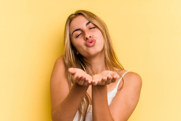 Junge kaukasische blonde Frau isoliert auf gelbem Hintergrund faltbare Lippen und Handflächen halten, um Luftkuss zu senden. - Foto, Bild