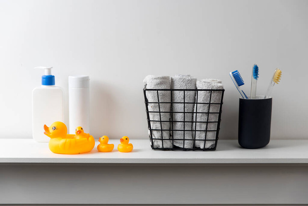 Shelf in the bathroom. Bouquet of gypsophila in white corrugated vase in style of the seventies, bottles of shampoo and cream, small face towels in container and yellow rubber ducks are on the shelf. - Foto, imagen