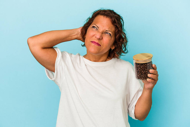 Middelbare leeftijd latijn vrouw met een koffiepot geïsoleerd op blauwe achtergrond raken achterkant van het hoofd, denken en het maken van een keuze. - Foto, afbeelding