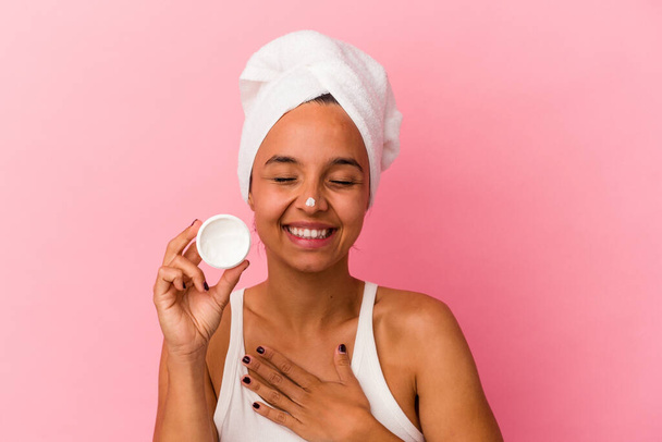 jong gemengd ras vrouw houden gezichtscrème geïsoleerd op roze achtergrond lachen en plezier hebben. - Foto, afbeelding